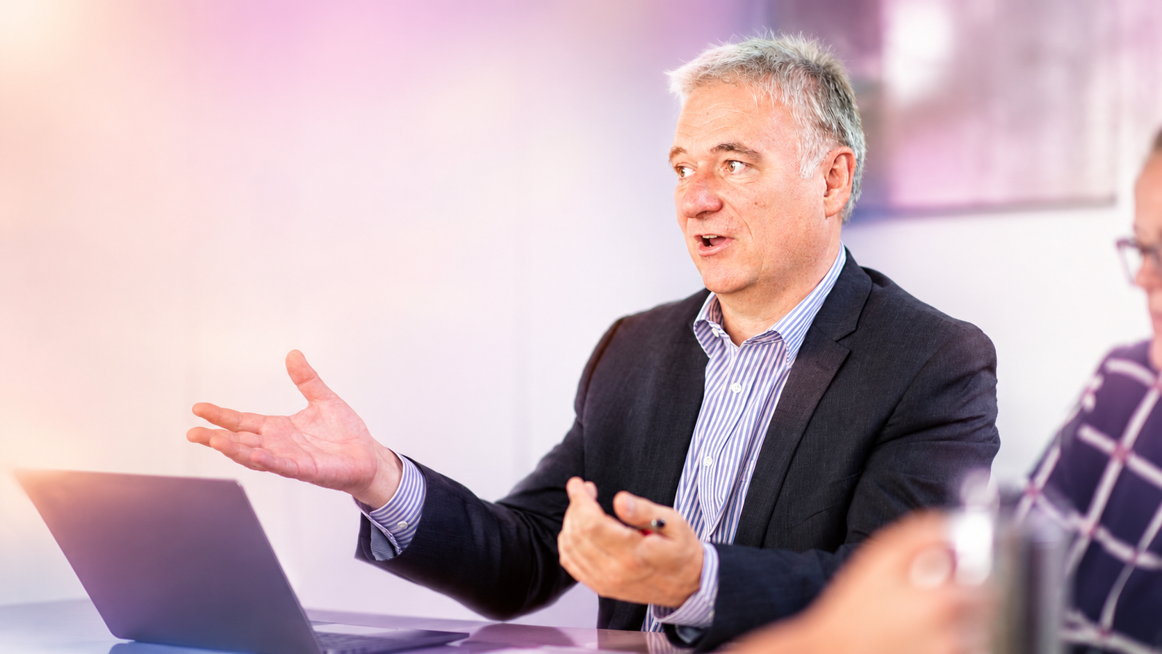 Man in a dark blazer and striped shirt speaks animatedly, gesturing with hands during a meeting with an open laptop on the table