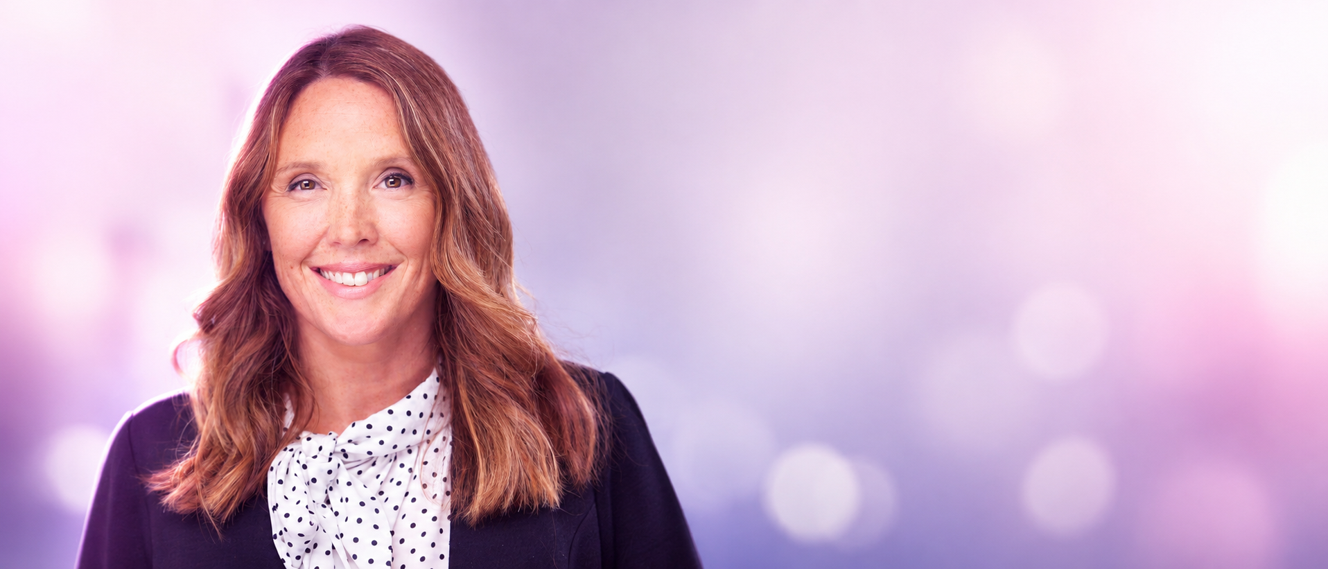 Smiling woman with light brown hair in a white polka-dot blouse and dark blazer, against a purple bokeh background.